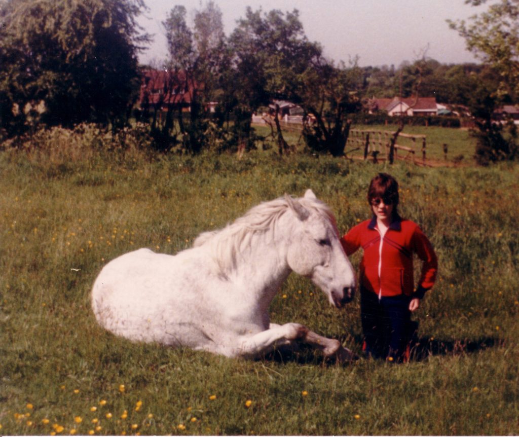 A grey pony is lying on the ground, snoozing. A girl in a 1980s red and blue tracksuit is kneeling next to him.